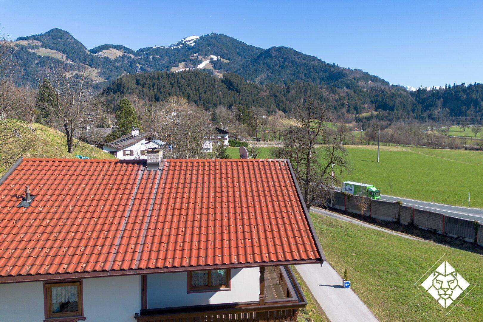 Blick über das rote Ziegeldach auf die umliegende Berglandschaft und grüne Felder.