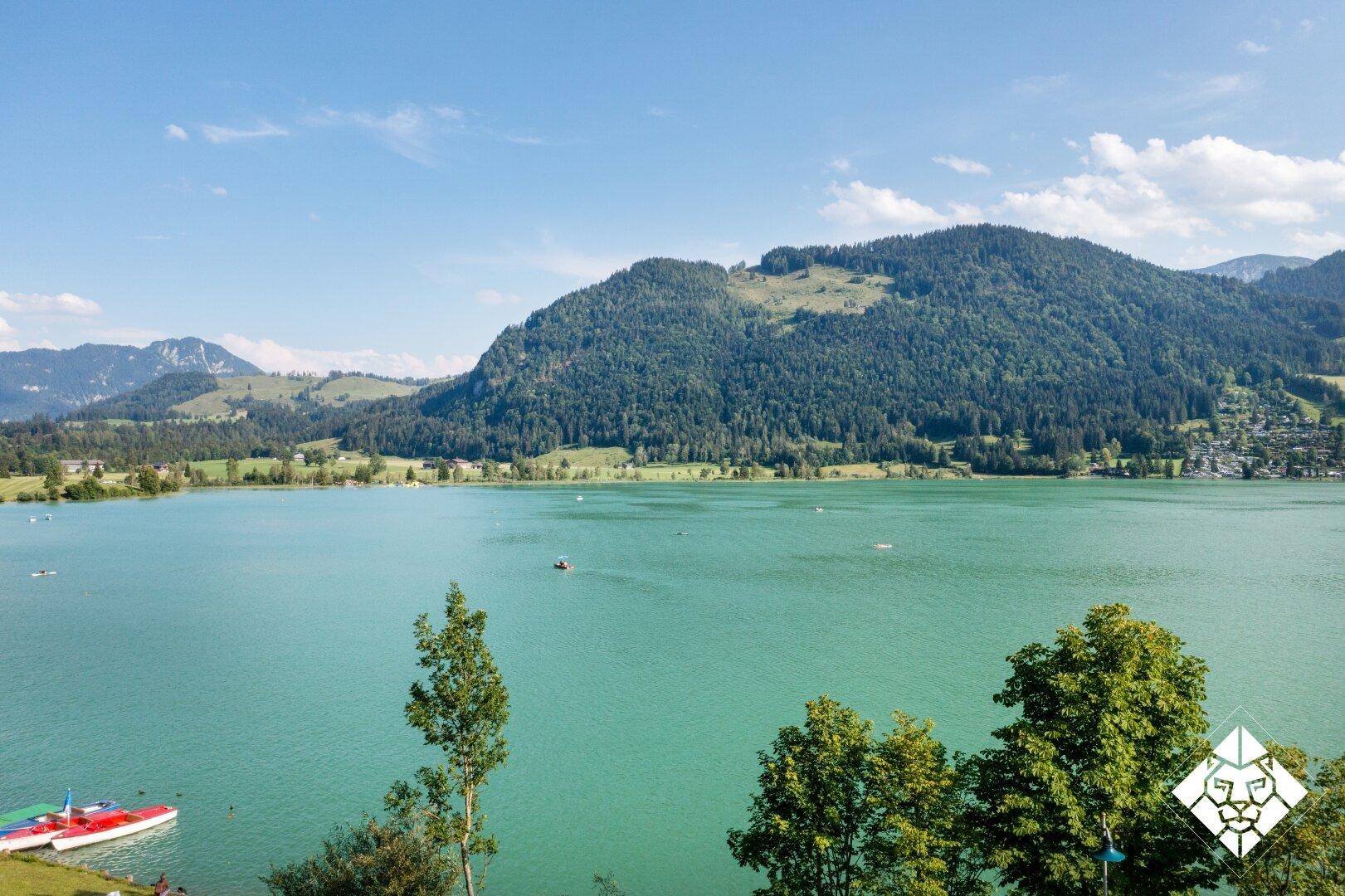 Panoramablick auf einen ruhigen See, umgeben von grünen Hügeln und majestätischen Bergen unter blauem Himmel.