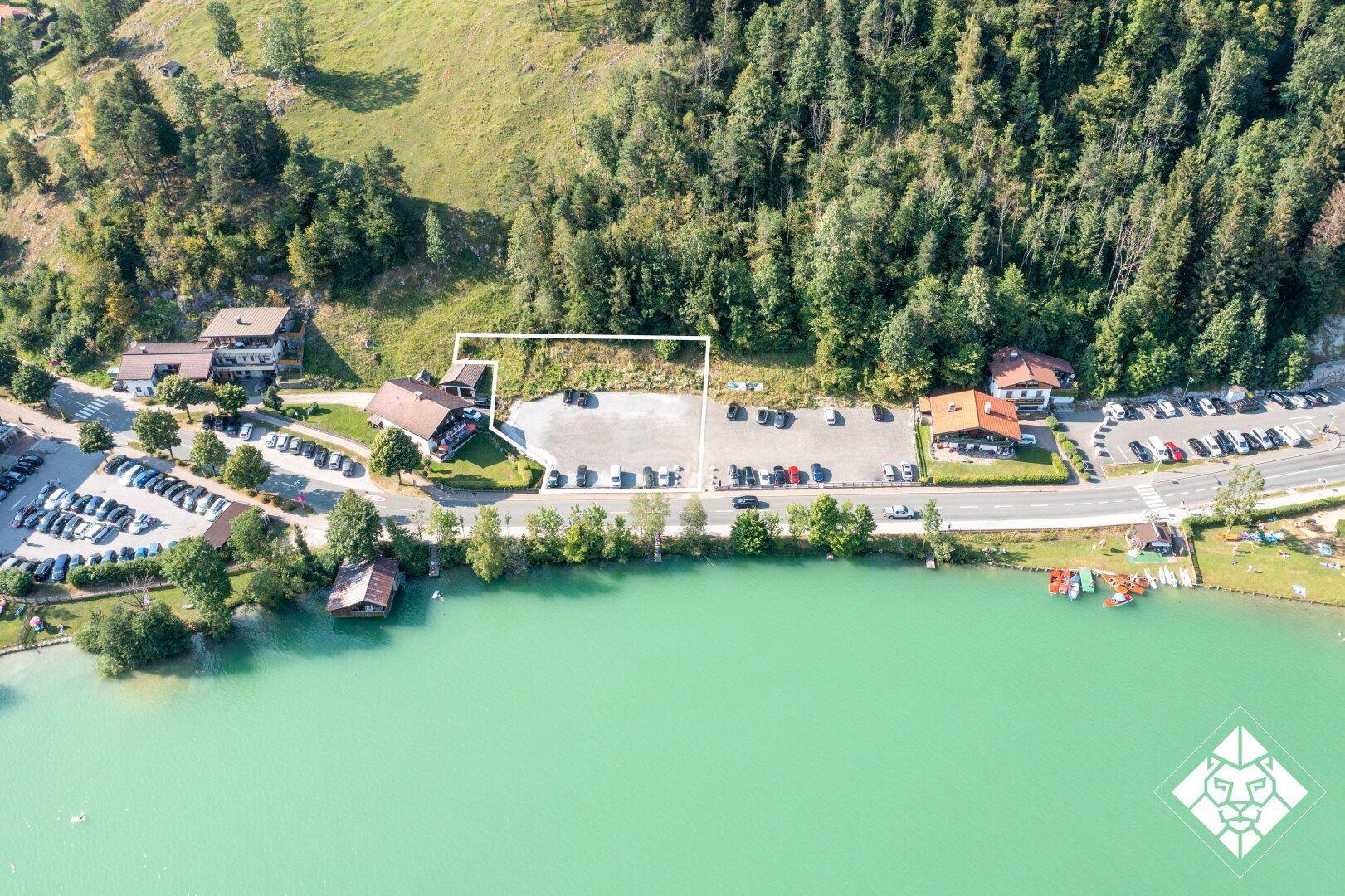 Luftaufnahme eines Grundstücks am Seeufer mit angrenzenden Parkplätzen und bewaldeten Hängen.