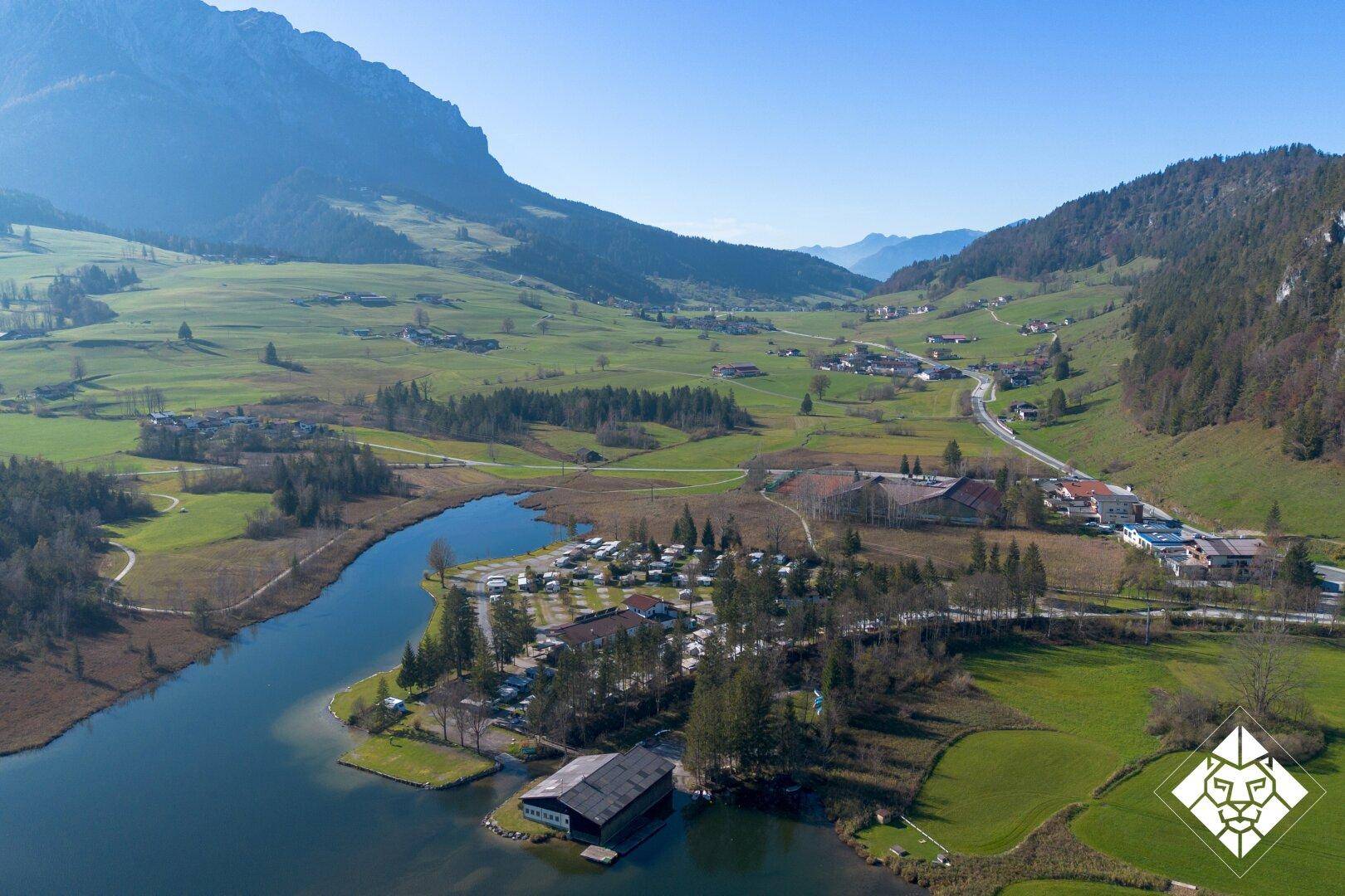 Luftaufnahme eines Tals mit Fluss, Campingplatz und umliegenden Bergen unter strahlend blauem Himmel.