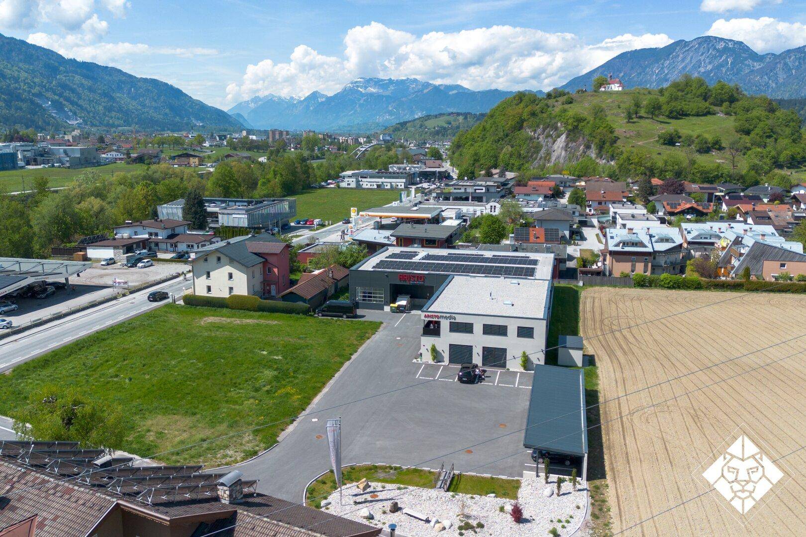 Luftaufnahme eines Gewerbegebäudes mit Parkplätzen, umgeben von grünen Feldern und majestätischen Bergen.