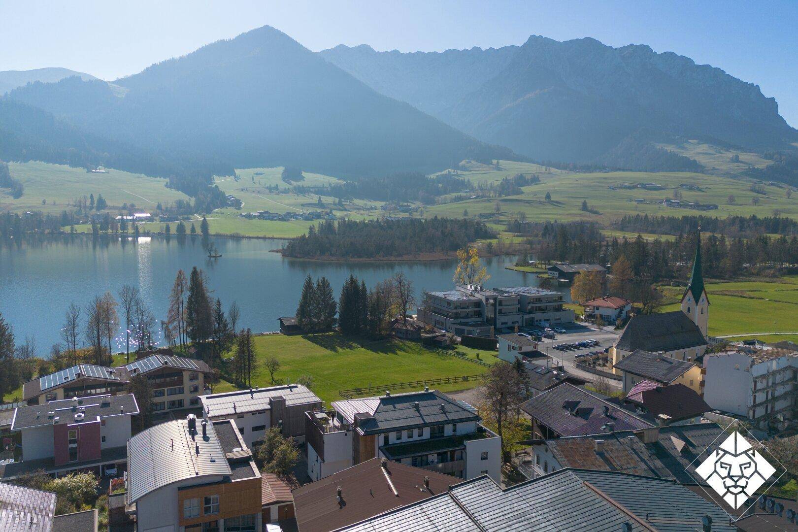 Panoramablick auf eine malerische Stadt am See, umgeben von grünen Hügeln und majestätischen Bergen.