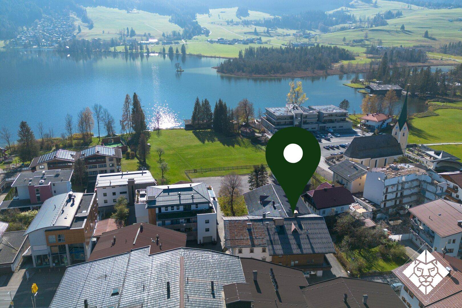 Luftaufnahme einer Siedlung am Seeufer mit einem Standort-Pin, umgeben von Natur und Bergen.