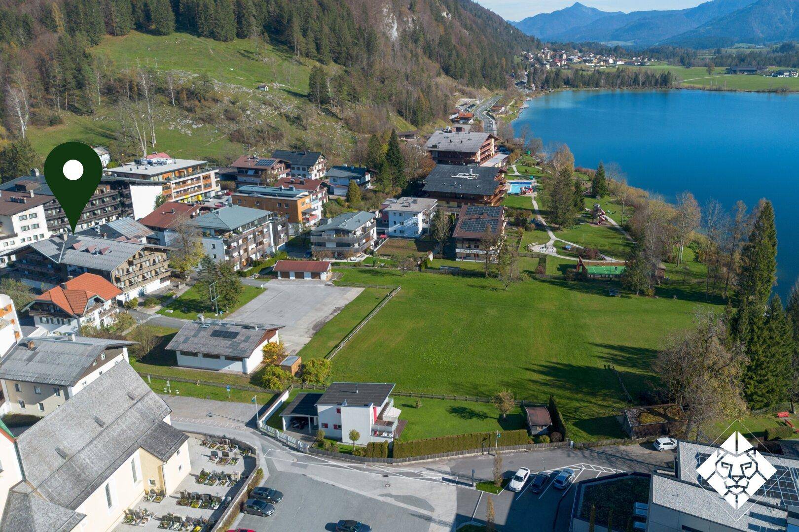 Luftaufnahme einer Stadt am Seeufer mit einem Standort-Pin, umgeben von grünen Wiesen und Bergen.