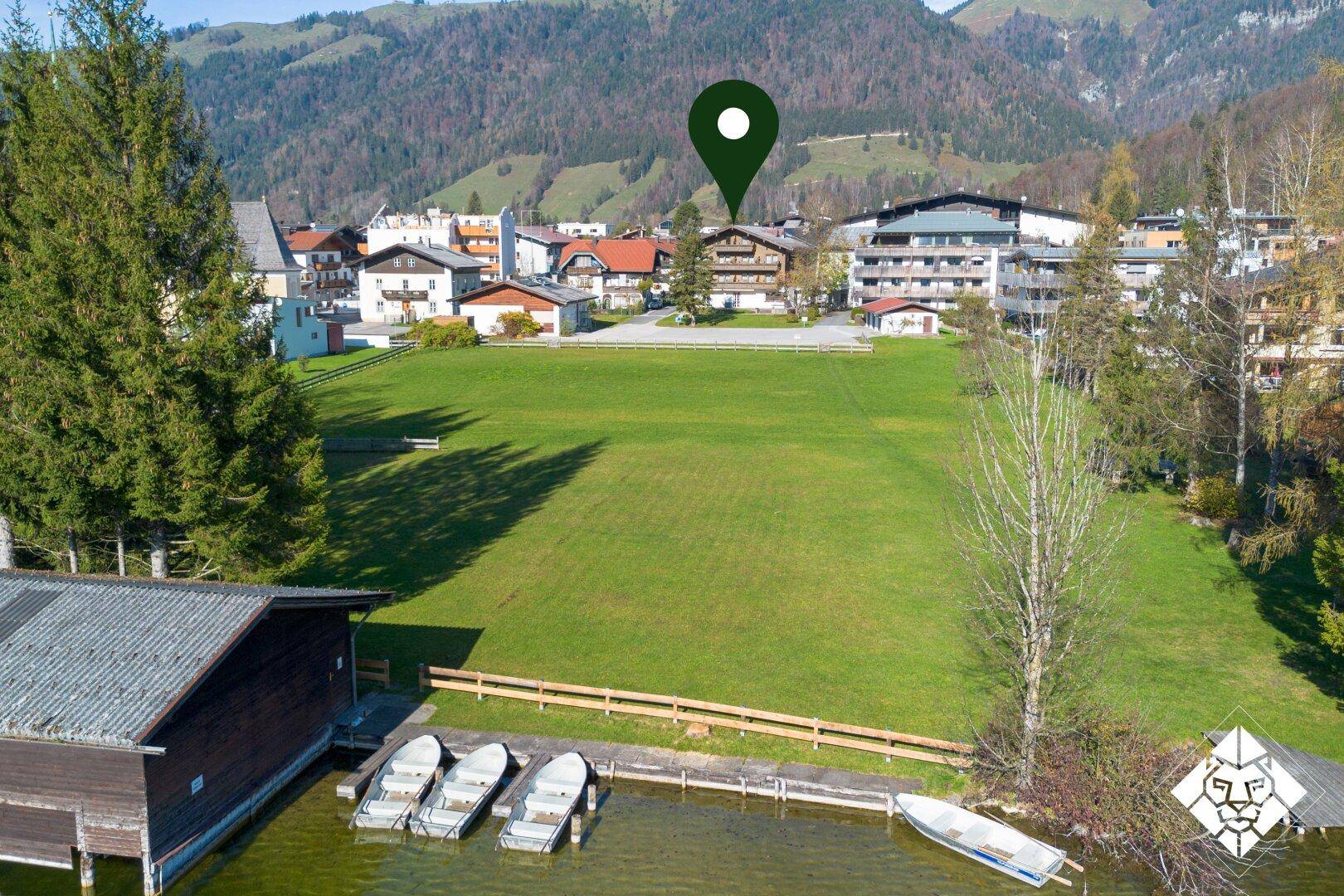 Luftaufnahme einer grünen Wiese am Seeufer mit Bootshaus und Booten, umgeben von Bergen.