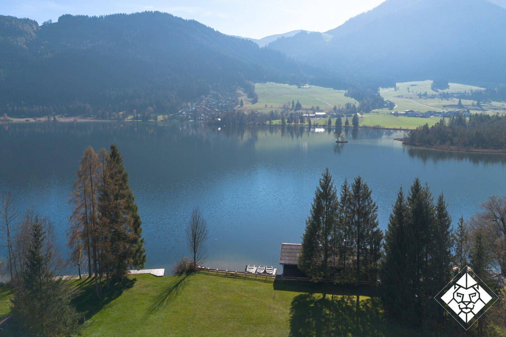 Luftaufnahme eines ruhigen Sees mit Bootshaus und grünen Wiesen, umrahmt von Bergen.