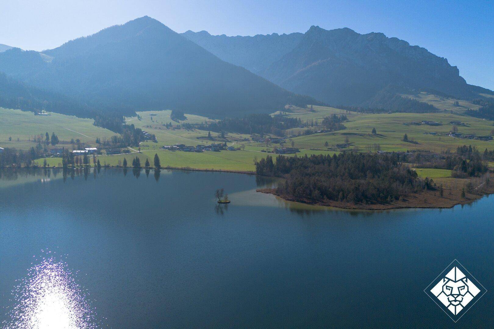 Weitläufige Luftaufnahme eines großen Sees mit kleiner Insel, umgeben von Bergen und grünen Feldern.