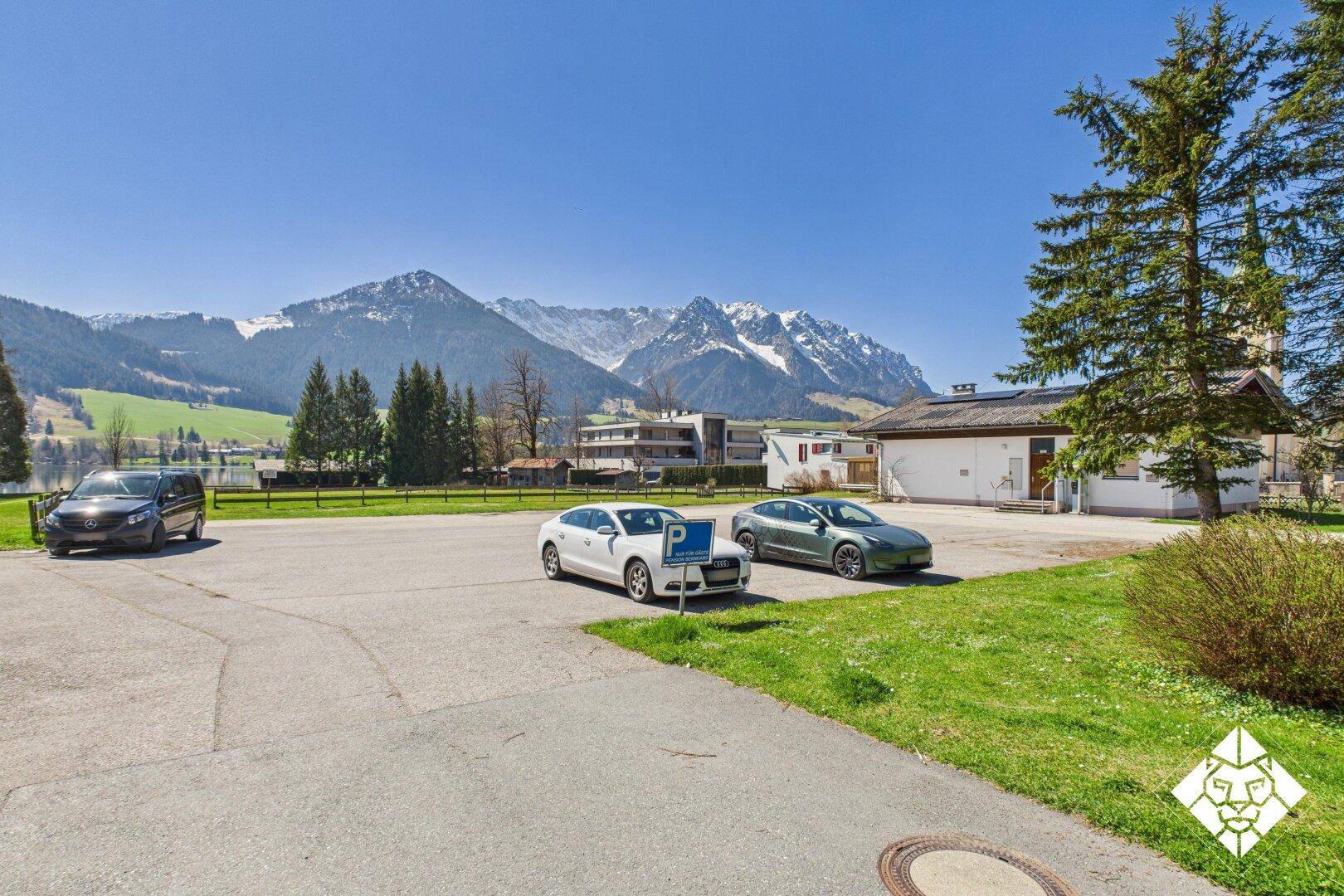 Außenparkplatz mit Autos, Bergen und See im Hintergrund unter klarem, blauem Himmel.