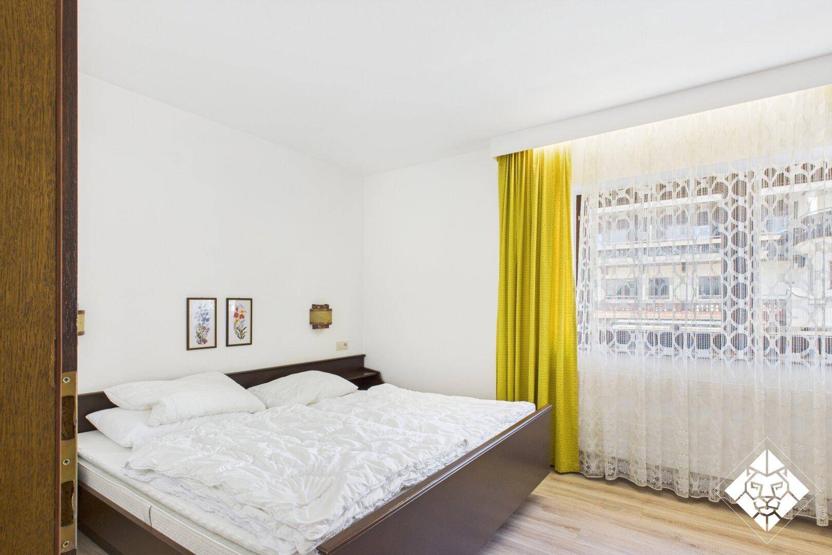 Schlafzimmer mit Doppelbett, dunklem Holzrahmen und gelben Vorhängen, mit Blick nach draußen.