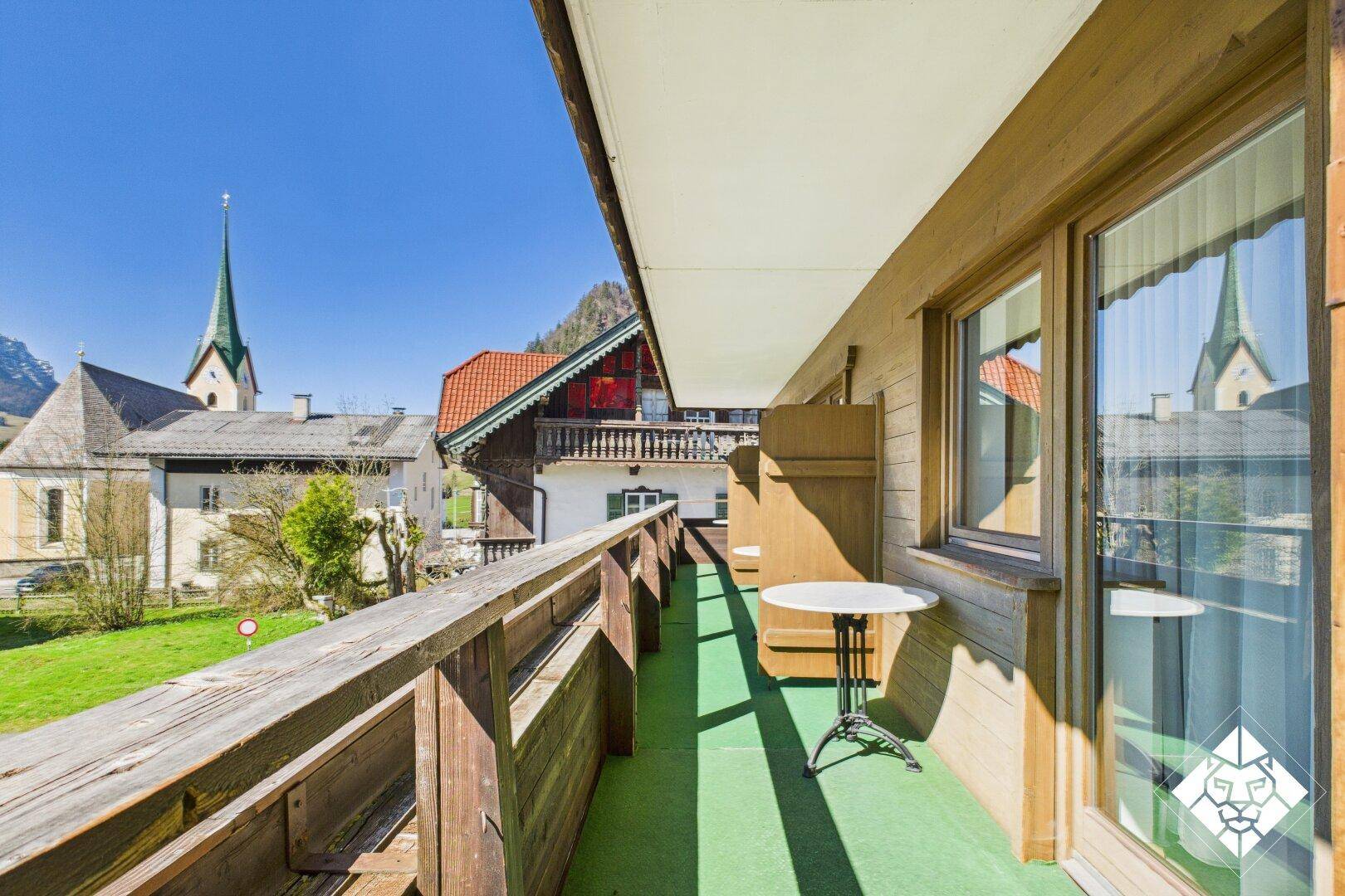 Langer Balkon mit Holzgeländer, grünem Teppich und kleinem Tisch, mit Blick auf eine Kirche.