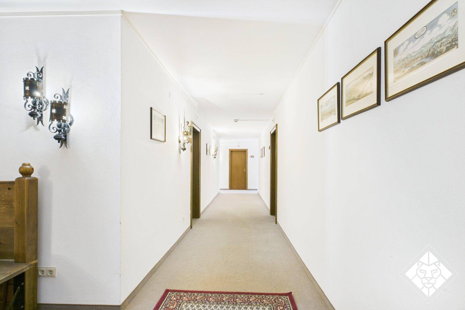 Langer Flur mit Teppichboden, weißen Wänden und Holztüren, beleuchtet von Wandleuchten.
