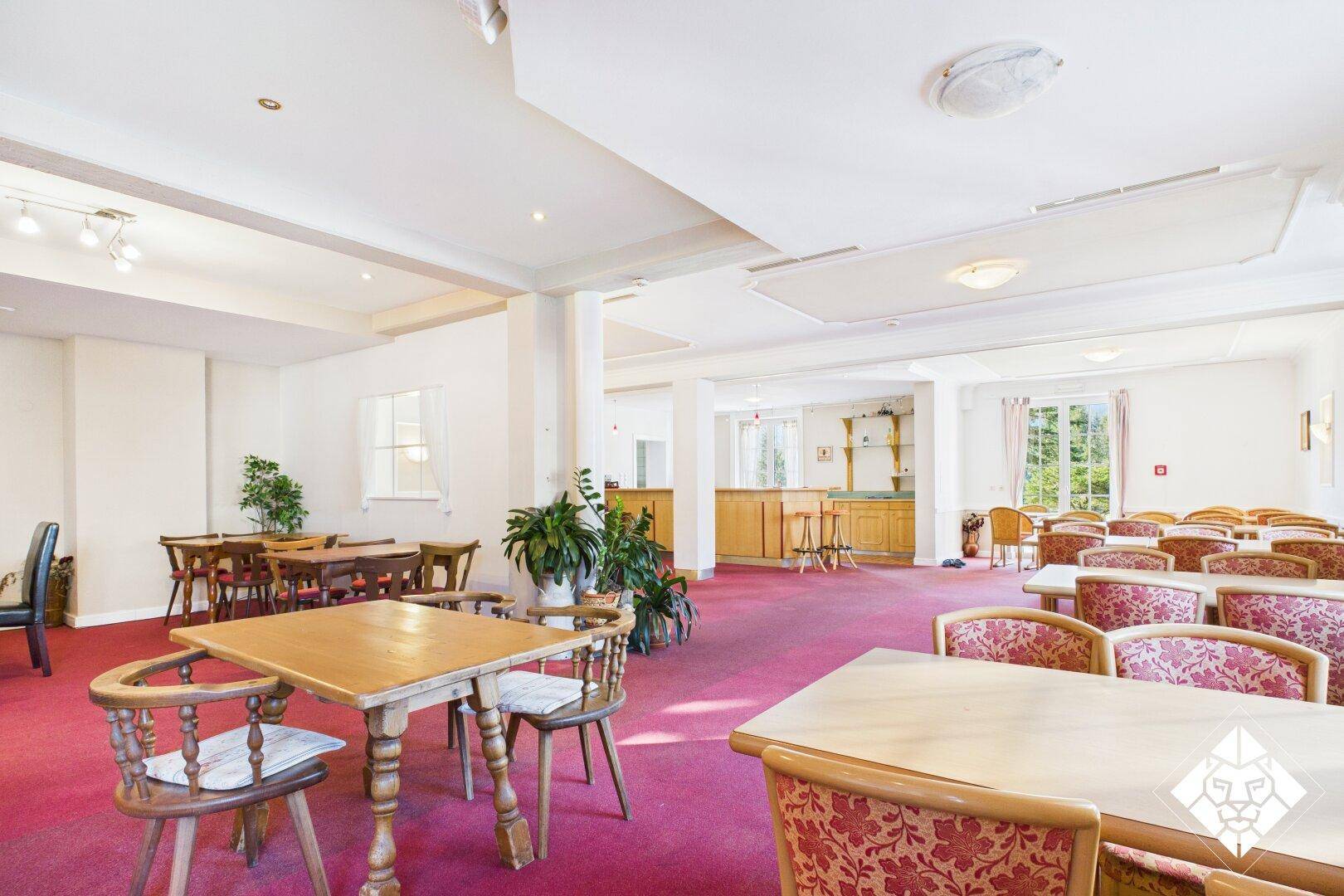 Großer Speisesaal mit vielen Tischen, roten Teppichböden und einer Bar im Hintergrund.