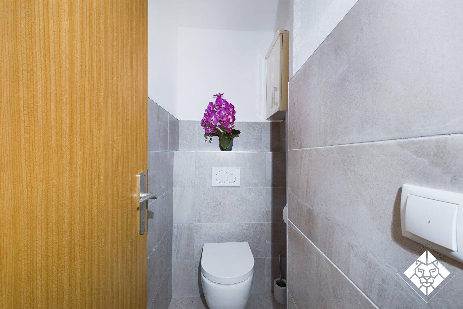 Modernes Gäste-WC mit wandhängender Toilette, grauen Fliesen und einer kleinen Ablage.