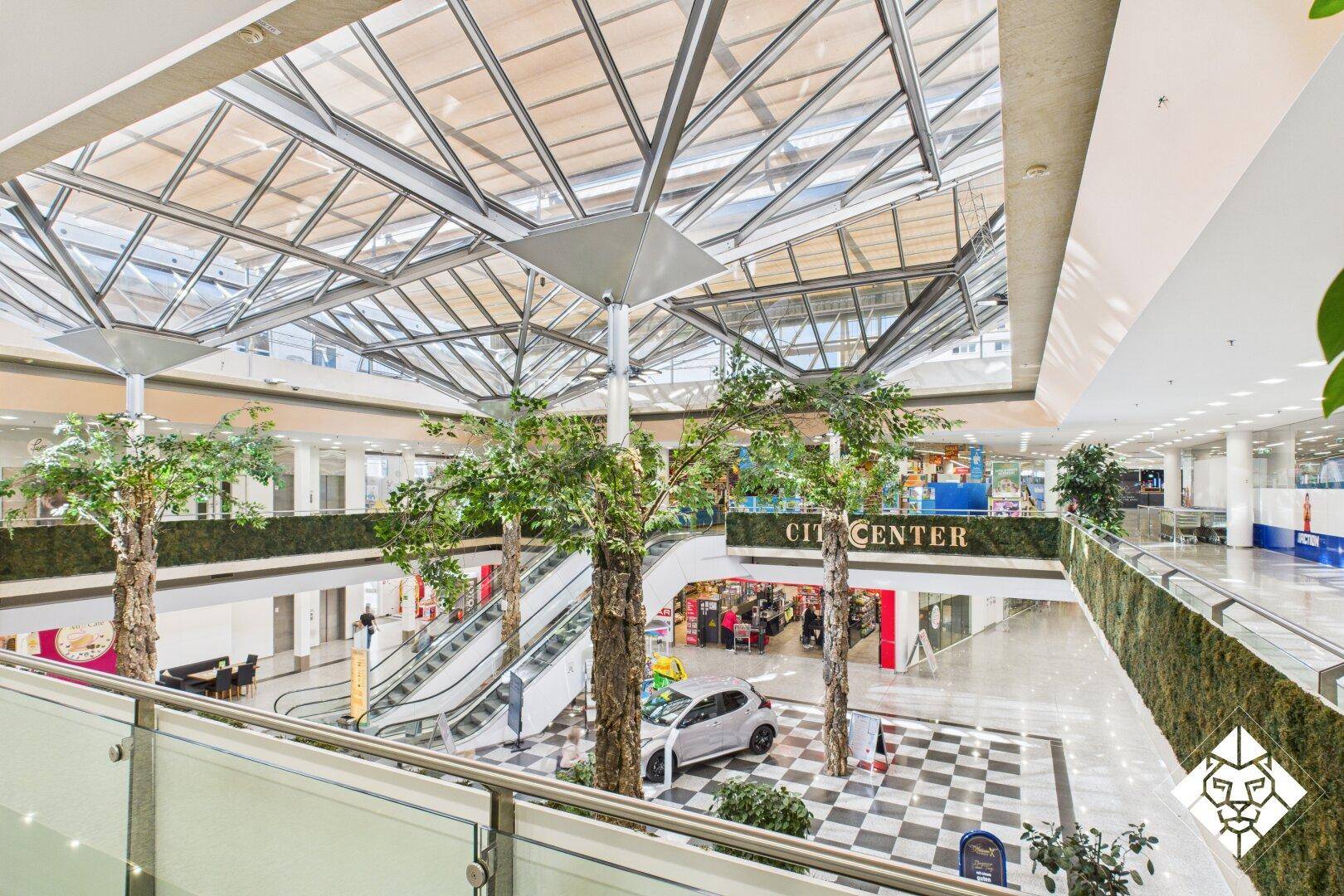 Innenansicht eines modernen Einkaufszentrums mit Glasdach, Rolltreppen und viel Tageslicht.