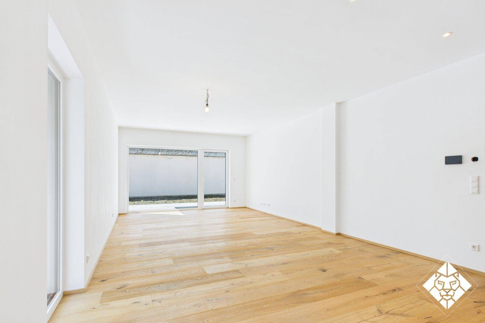 Helles, leeres Wohnzimmer mit Holzboden, großen Fenstern und Zugang zum Außenbereich.