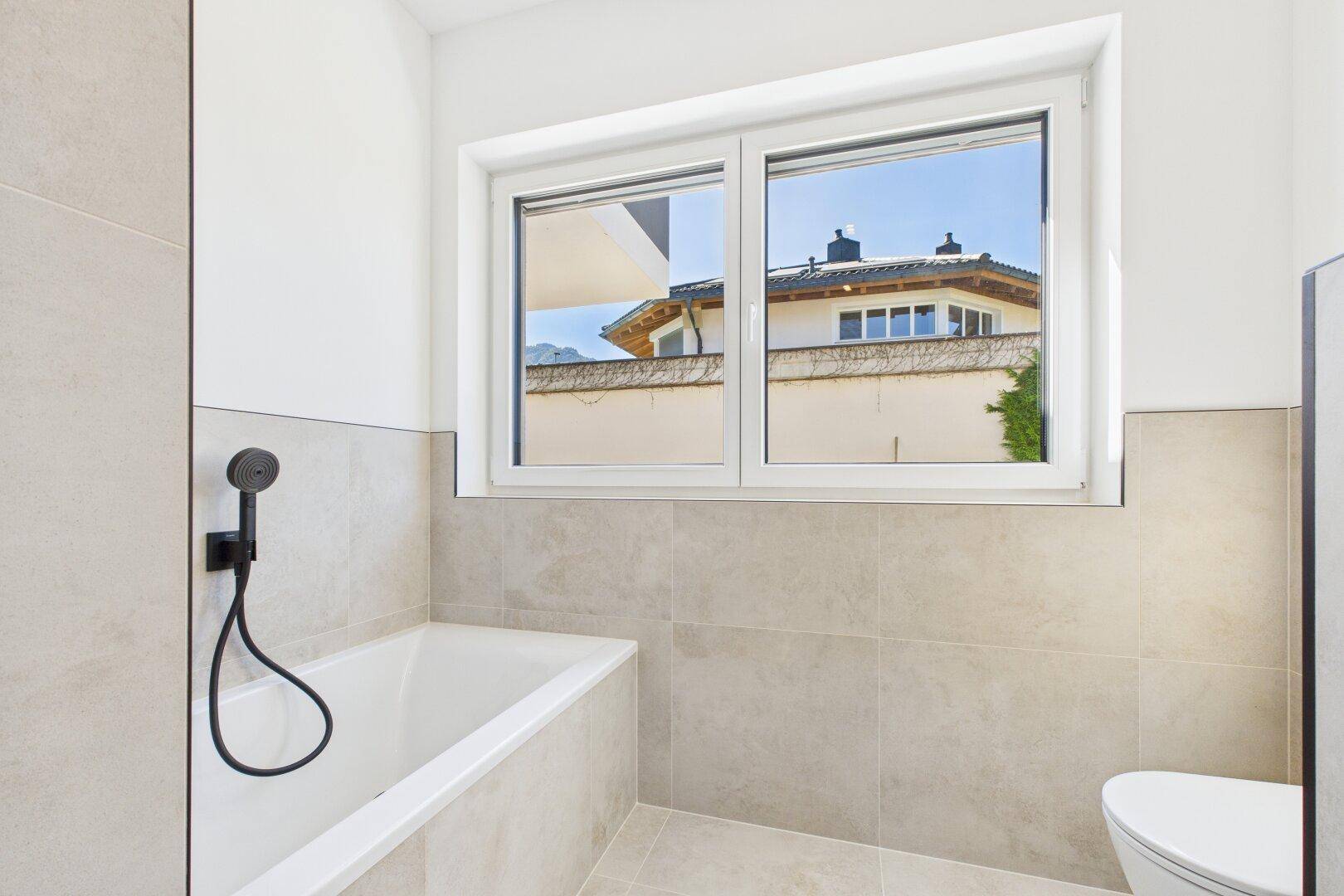 Helles Badezimmer mit Badewanne, Fenster und moderner Toilette, alles in neutralen Tönen.