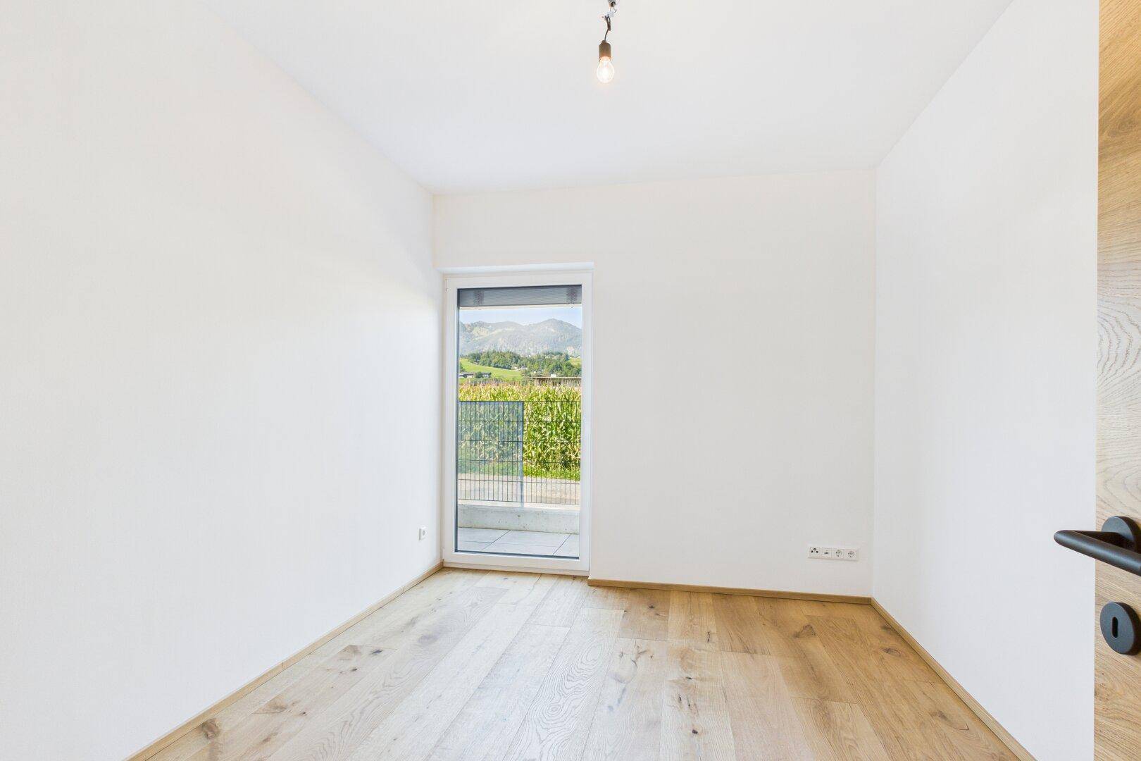 Helles Schlafzimmer mit Holzboden und Fenstertür, die auf einen Balkon oder eine Terrasse führt.