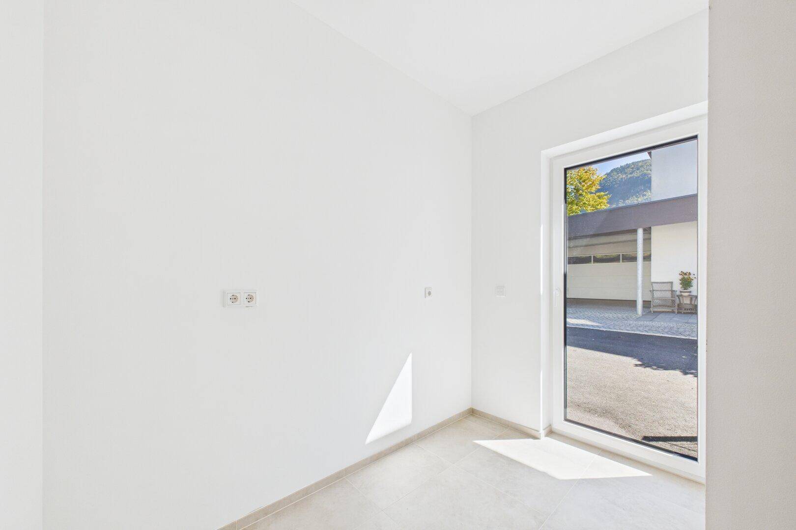 Heller Raum mit Fliesenboden und großer Fenstertür, die nach draußen führt.