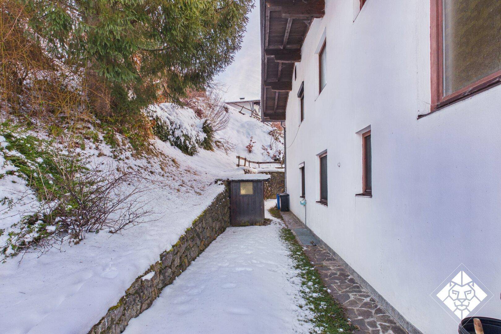 Schmaler Gartenweg entlang des Hauses mit Steinmauer und schneebedeckter Böschung.