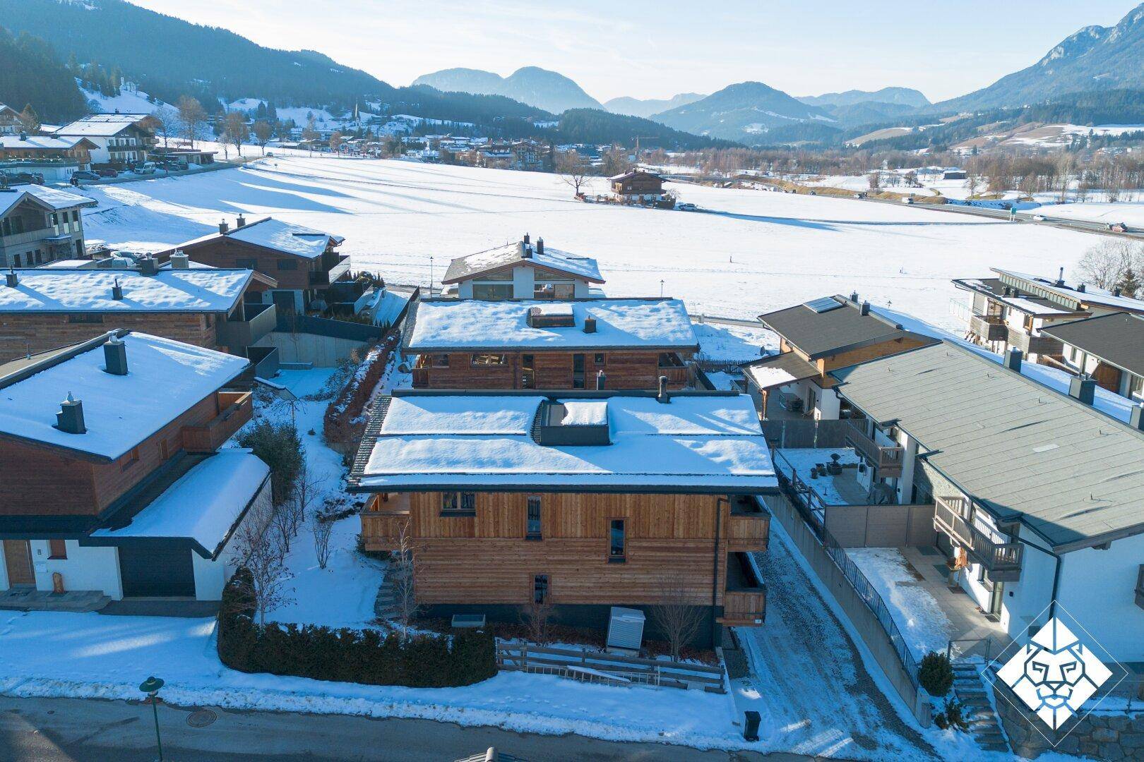 Luftaufnahme einer Gruppe von Holzchalets in einer verschneiten Berglandschaft mit weiten Feldern.