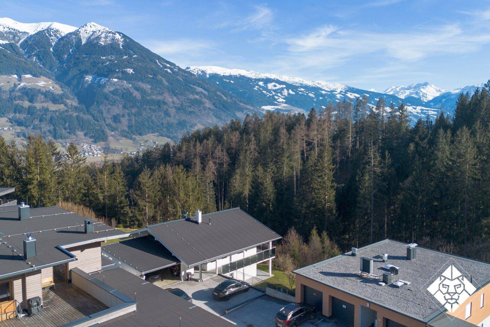 Luftaufnahme des Hauses in alpiner Umgebung mit bewaldeten Hängen und schneebedeckten Bergen.