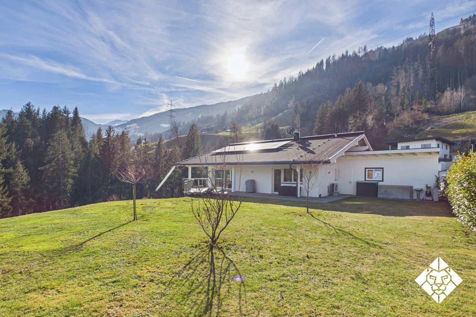 Haus in sonniger Hanglage mit weitläufigem Rasen und Blick auf die umliegende Berglandschaft.