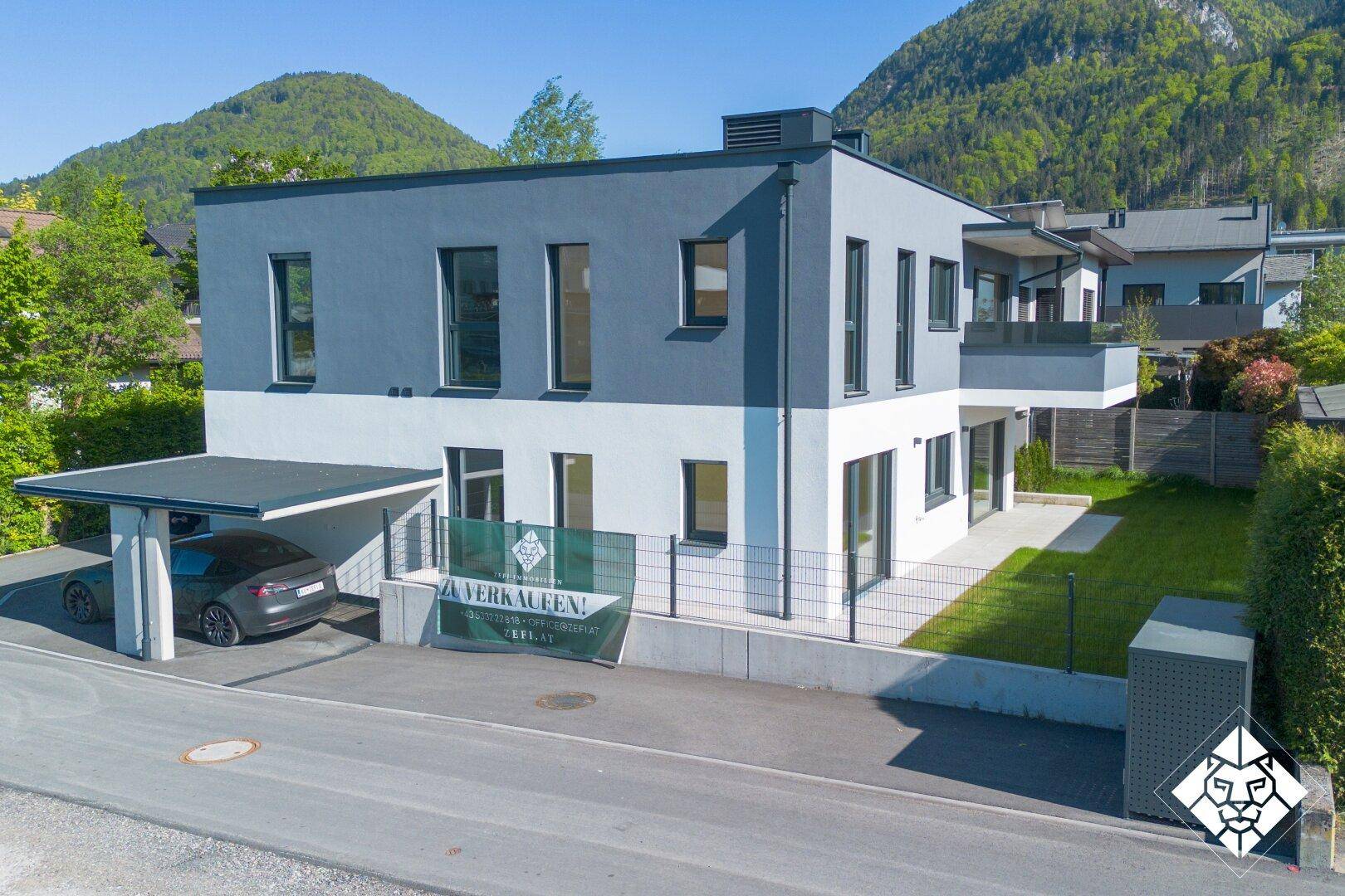 Modernes Haus mit Carport und gepflegtem Garten, umgeben von einer malerischen Berglandschaft.