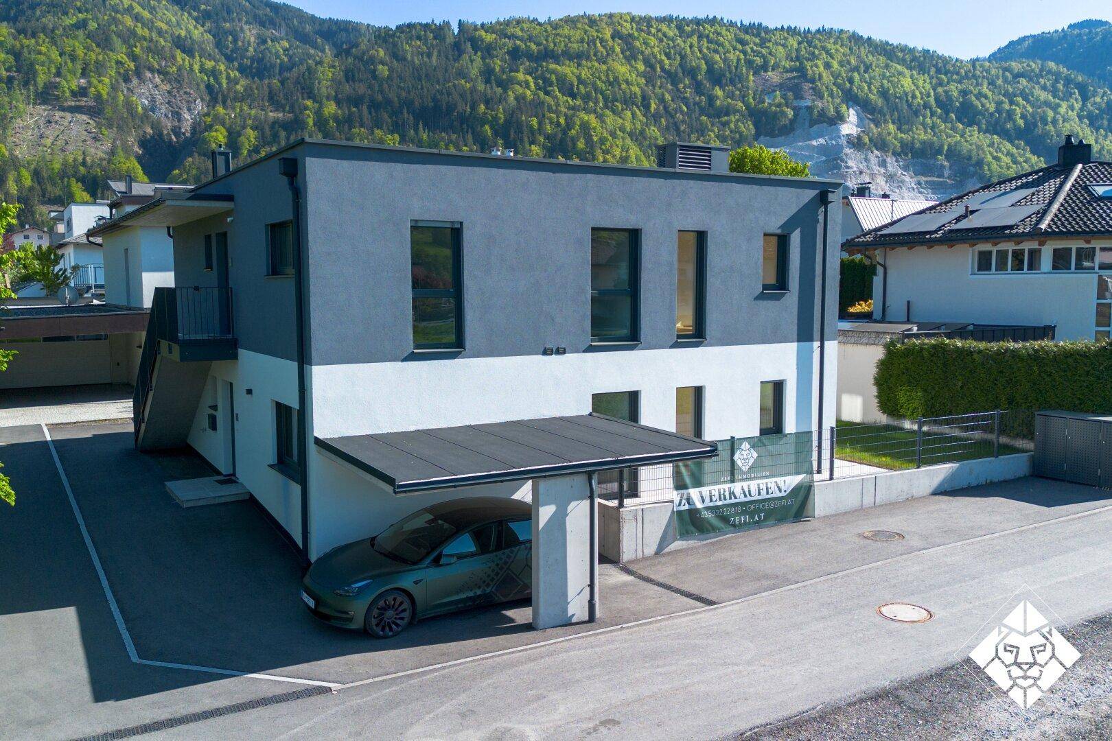 Ansicht des modernen Hauses mit Carport und Zufahrt, eingebettet in eine grüne Umgebung mit Bergblick.