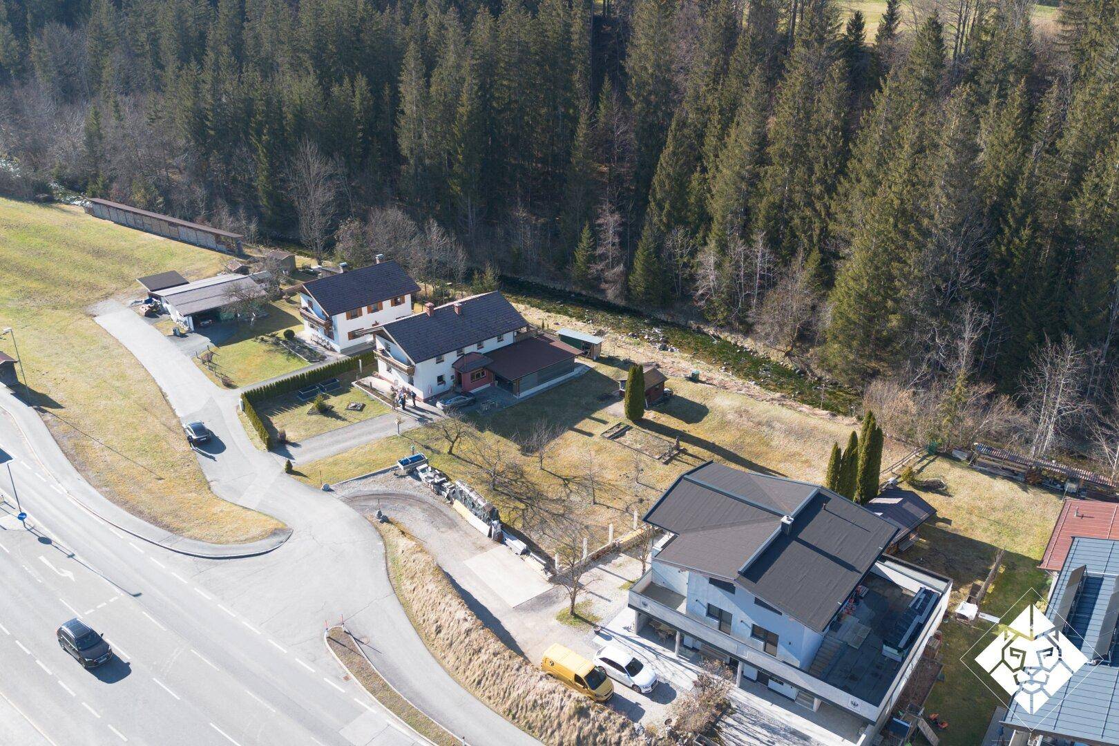 Weitläufige Luftaufnahme einer Wohnsiedlung mit Wald, Straße und Bachlauf in ländlicher Umgebung.