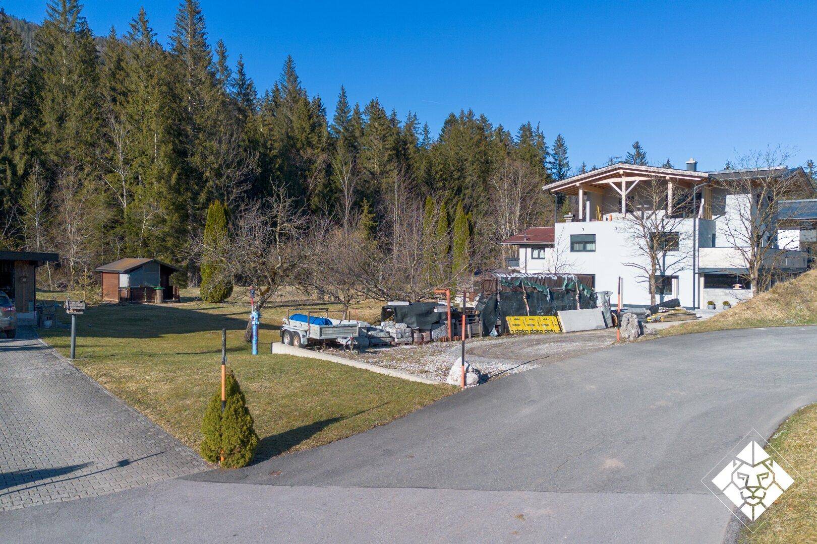 Ansicht eines im Bau befindlichen Hauses mit Rohbau, gepflasterter Zufahrt und Grünfläche im Vordergrund.
