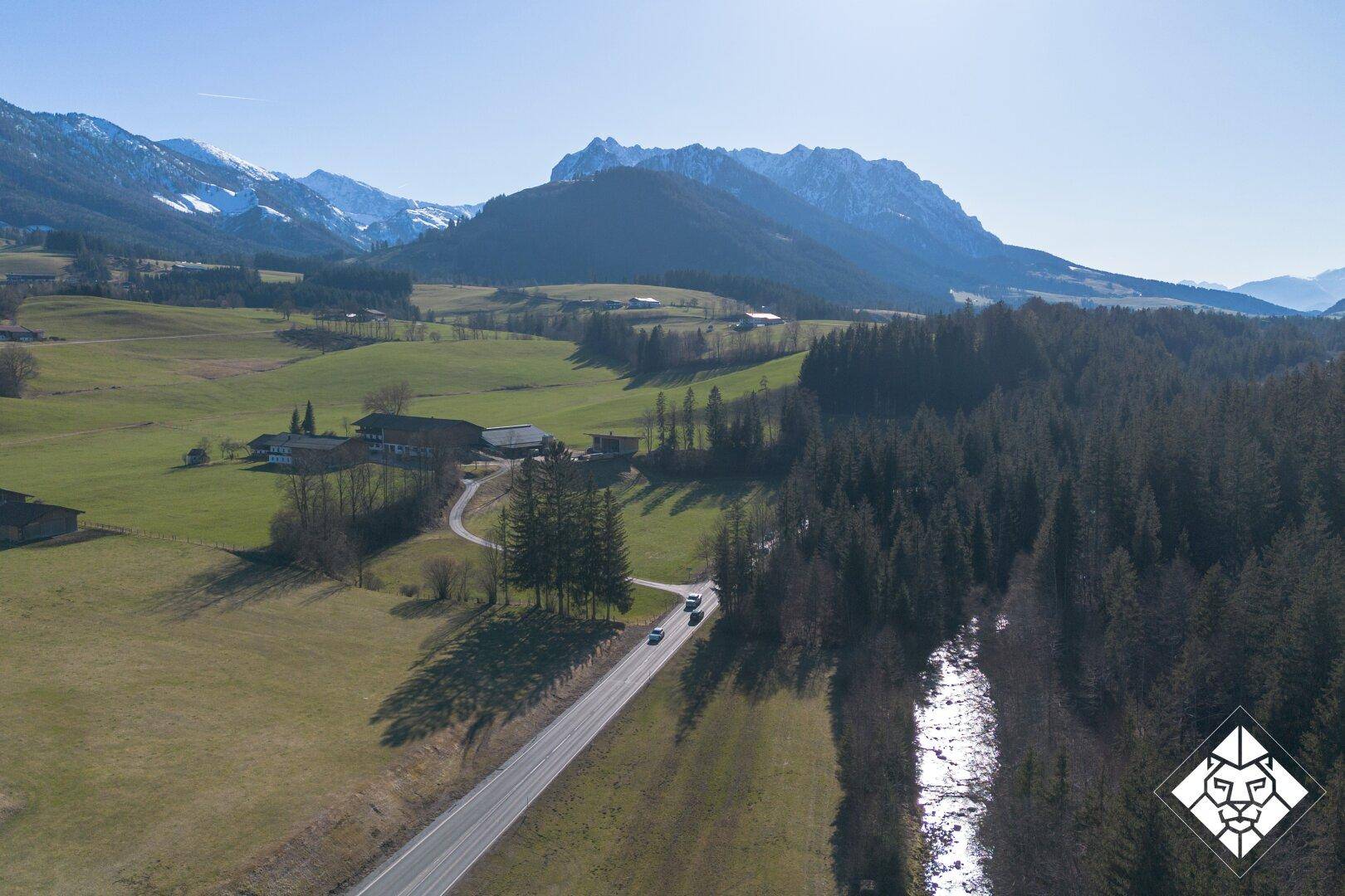 Malerische Luftaufnahme eines Tals mit Straße, Wald, Wiesen und schneebedeckten Bergen im Hintergrund.