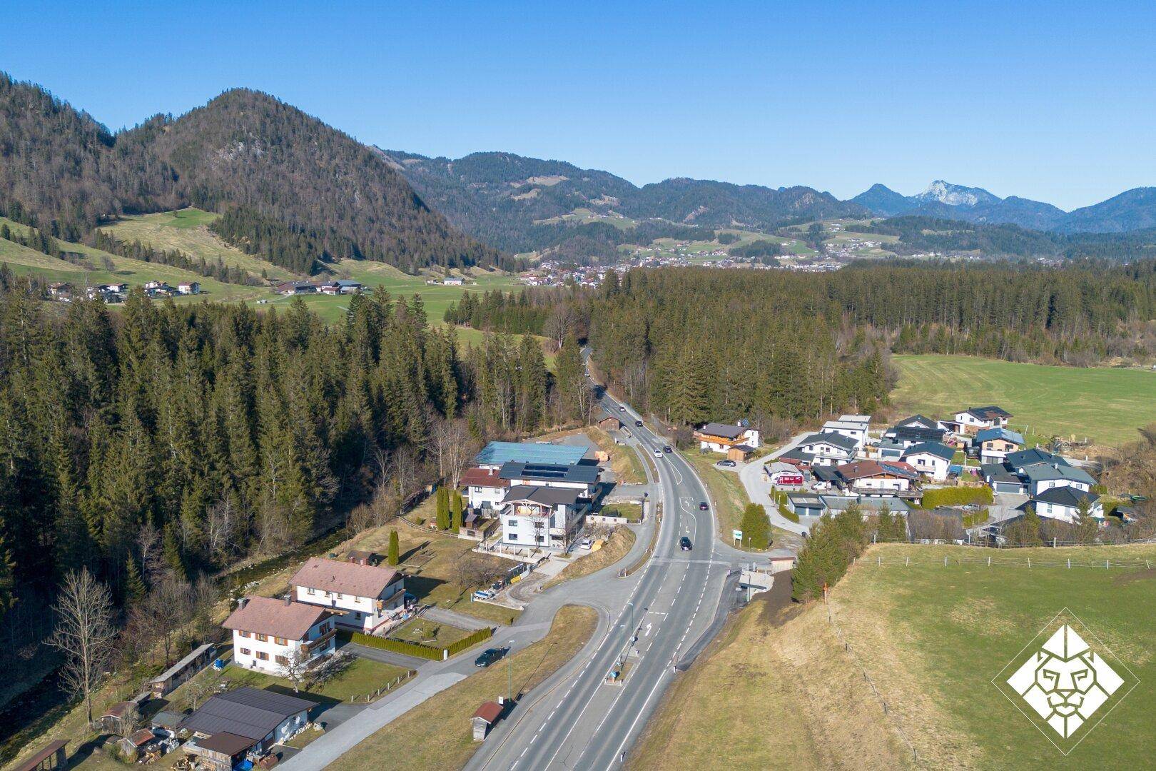 Weitwinkel-Luftaufnahme einer ländlichen Ortschaft, umgeben von Wäldern, Bergen und einer Hauptstraße.