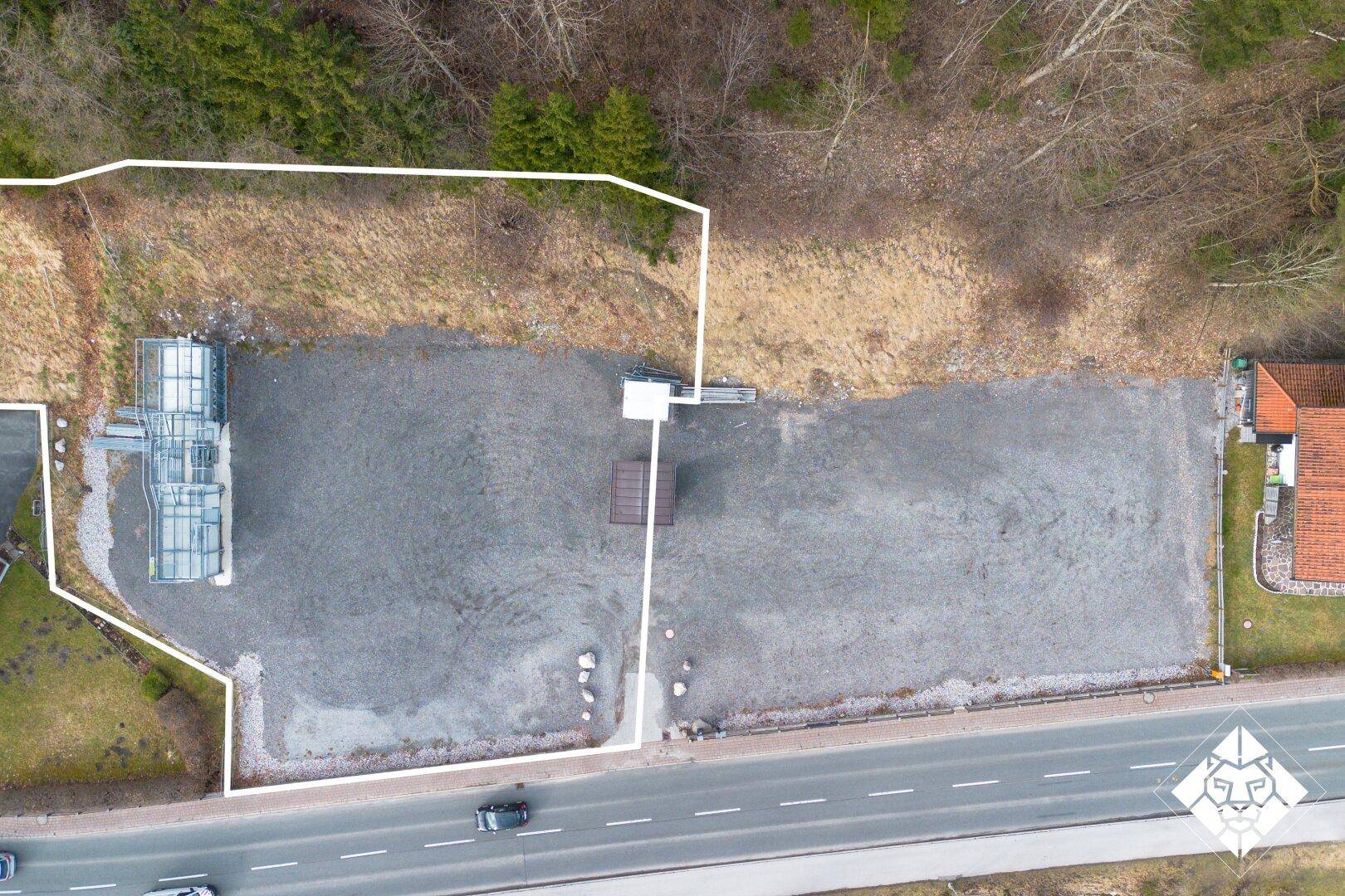 Luftaufnahme eines Kiesgrundstücks an einer Straße mit einem modernen Gebäude und Wald im Hintergrund.