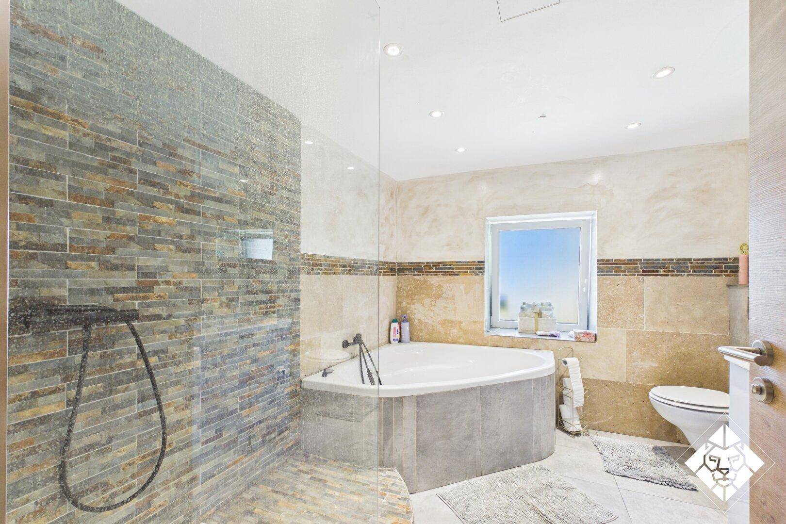 Modernes Badezimmer mit einer Eckbadewanne, einer begehbaren Dusche und Fliesen in Natursteinoptik.