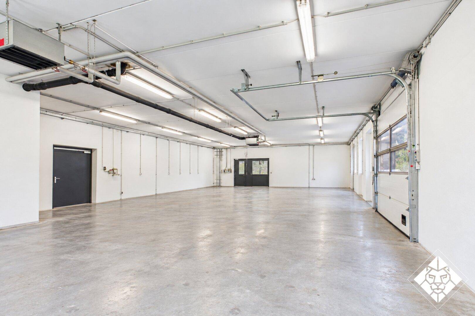 Geräumiger Lagerraum mit Betonboden, Rolltor, industrieller Deckeninstallation und Fenstern.