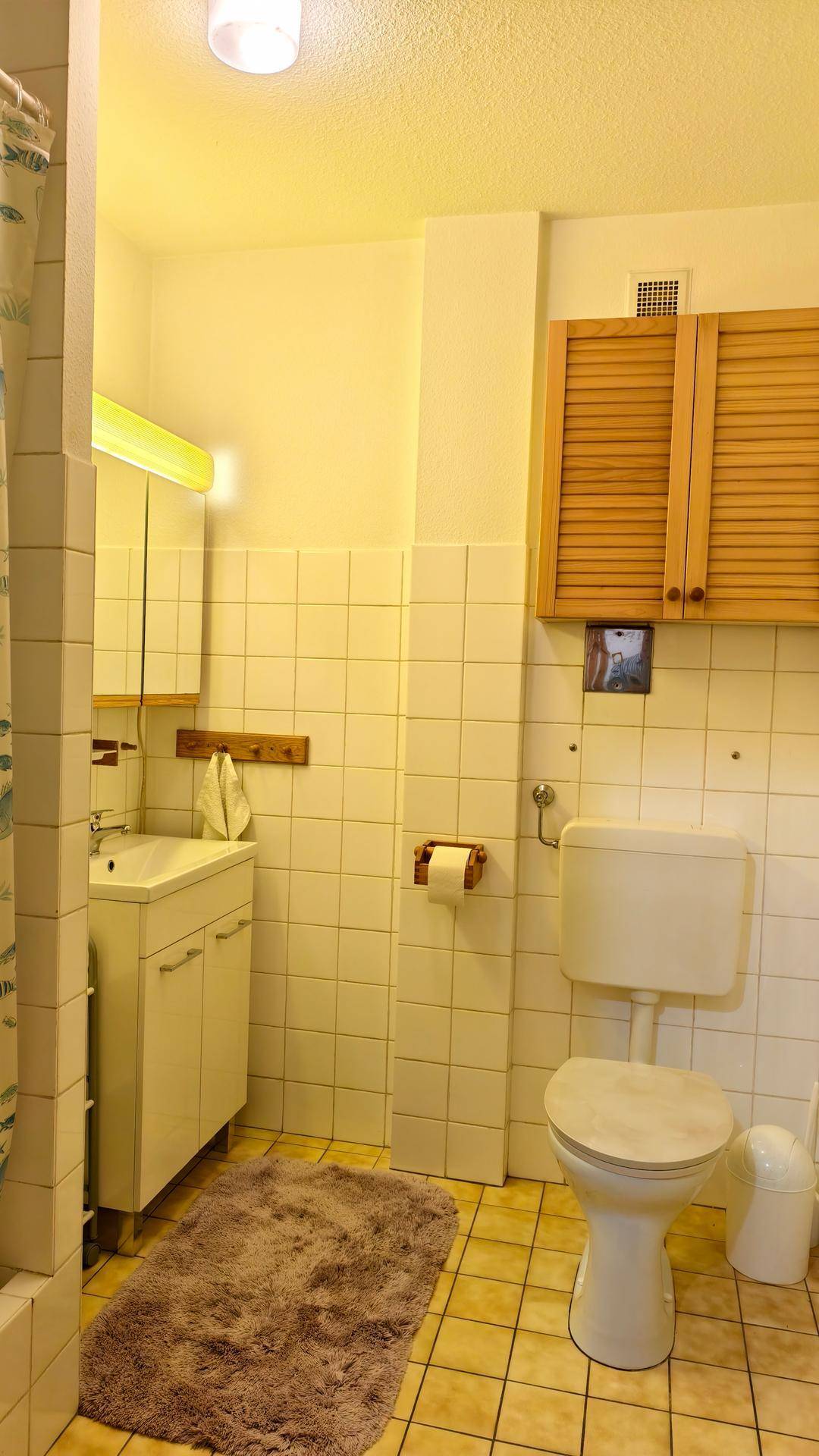 Badezimmer mit weißen Fliesen, Toilette, Waschbecken und Spiegelschrank unter gelblichem Licht.