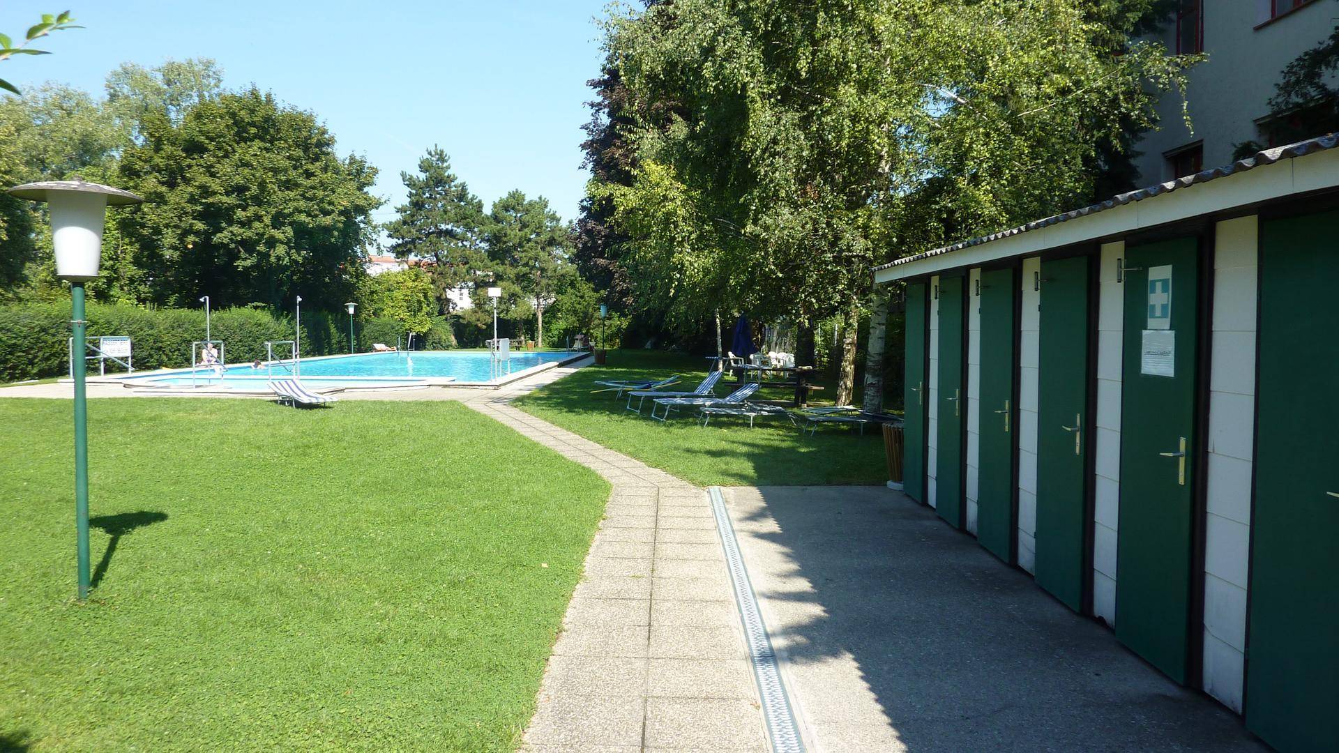 Außenbereich mit Schwimmbecken, Liegewiese und Umkleidekabinen unter blauem Himmel.