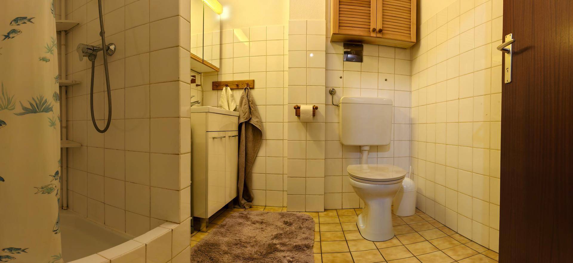 Gefliestes Badezimmer mit Duschbereich, Toilette, Waschbecken und Holzschrank über dem WC.