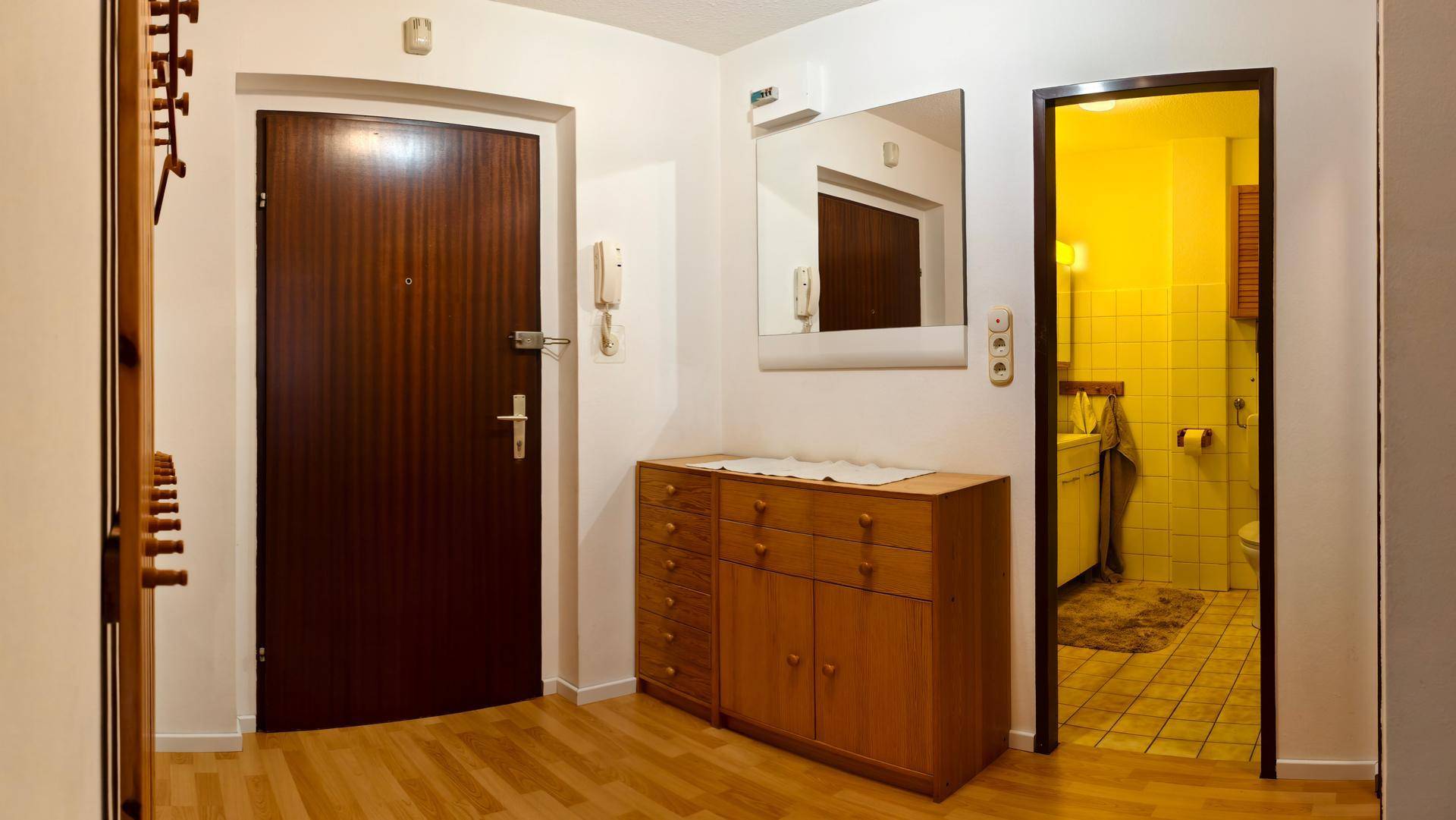 Eingangsbereich mit dunkler Haustür, Holzkommode und Spiegel, Zugang zum Badezimmer.