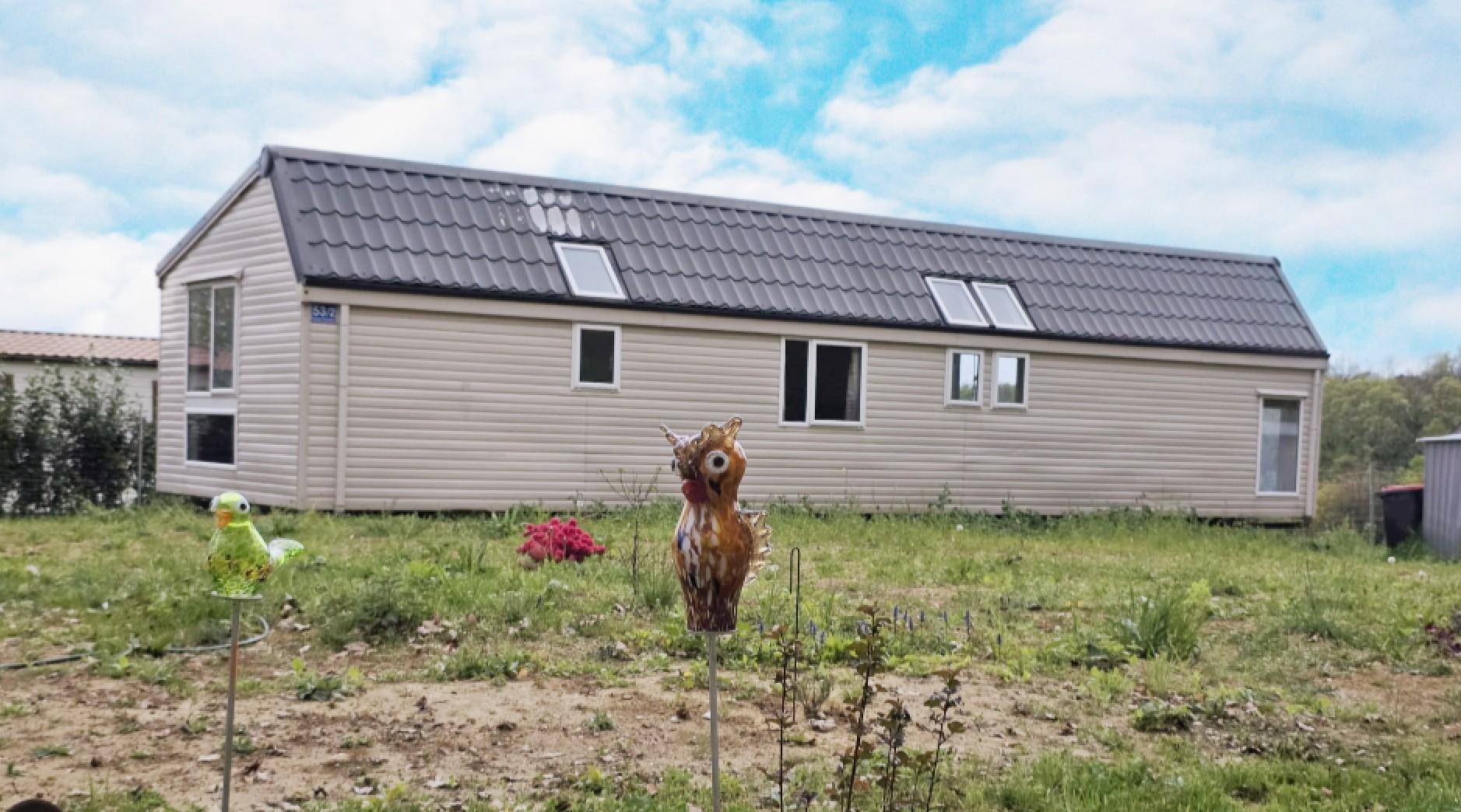Langes Mobilheim mit dunklem Satteldach und mehreren Dachfenstern in ländlicher Umgebung mit Wiese.