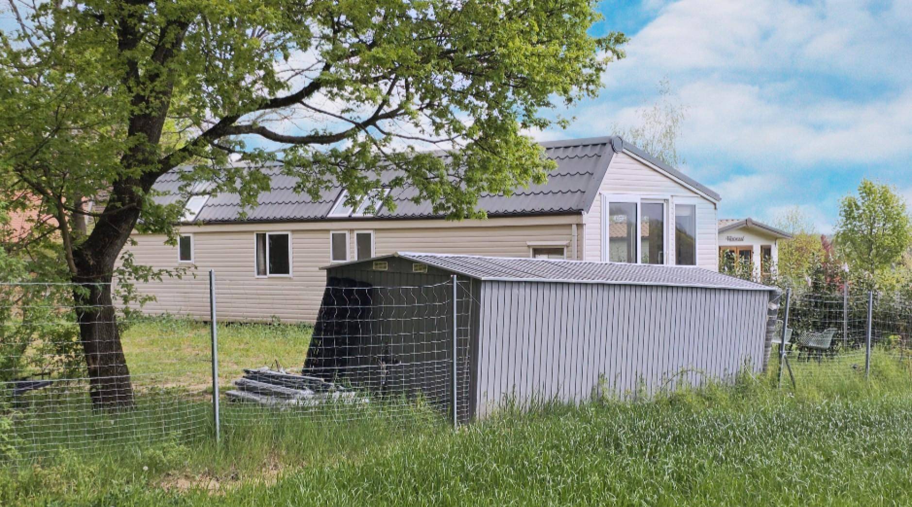 Mobilheim im Hintergrund mit einem Gartenhaus und einem Baum im Vordergrund, umgeben von Grünflächen.