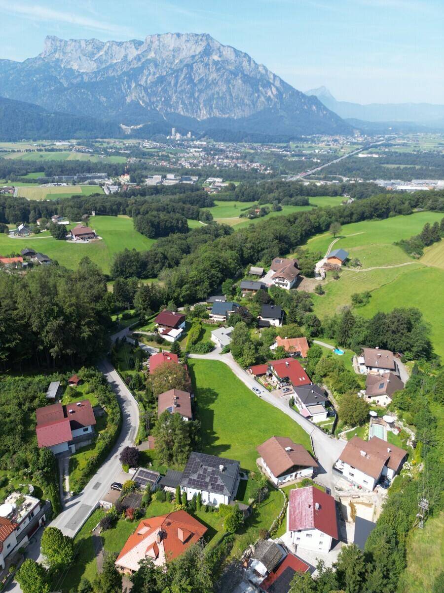 Weitläufige Ansicht einer ländlichen Siedlung mit beeindruckendem Bergpanorama und urbaner Silhouette am Horizont.