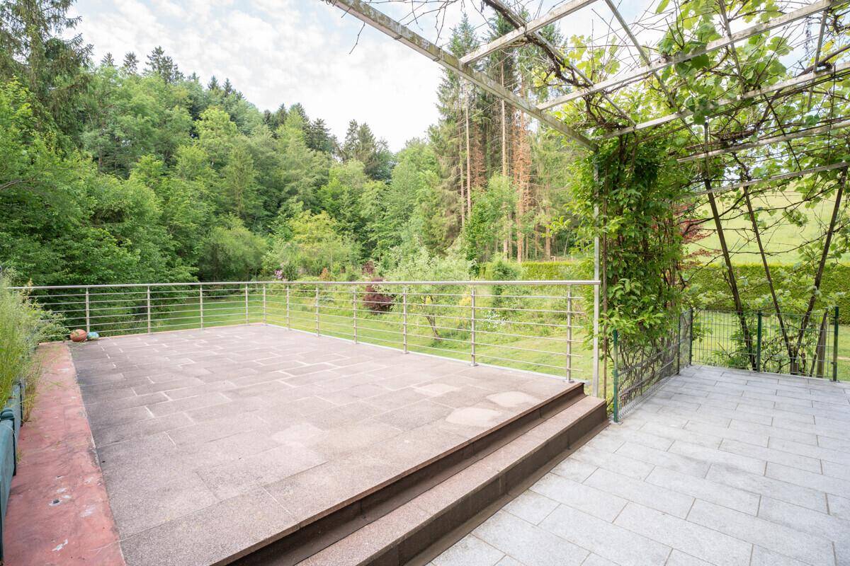Große gepflasterte Terrasse mit Pergola und Geländer, umgeben von viel Grün.