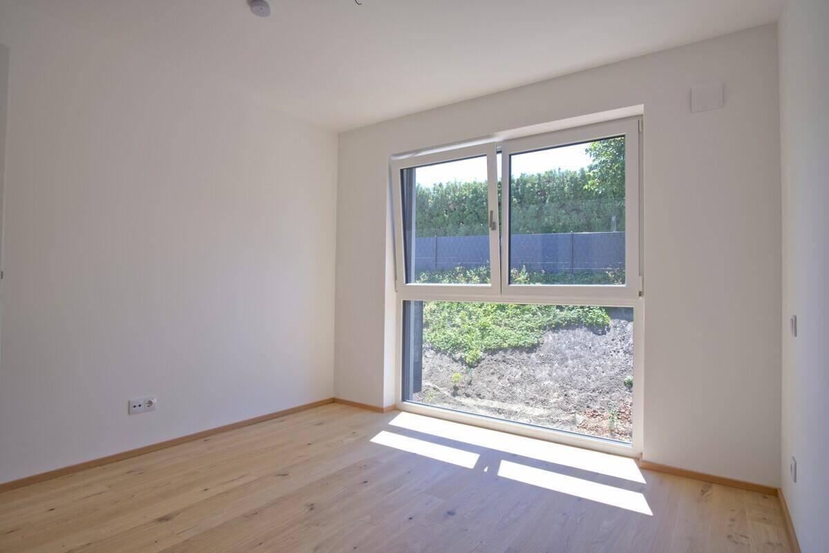 Sonniges Schlafzimmer mit hellem Parkettboden und großem Fenster mit Blick ins Grüne.