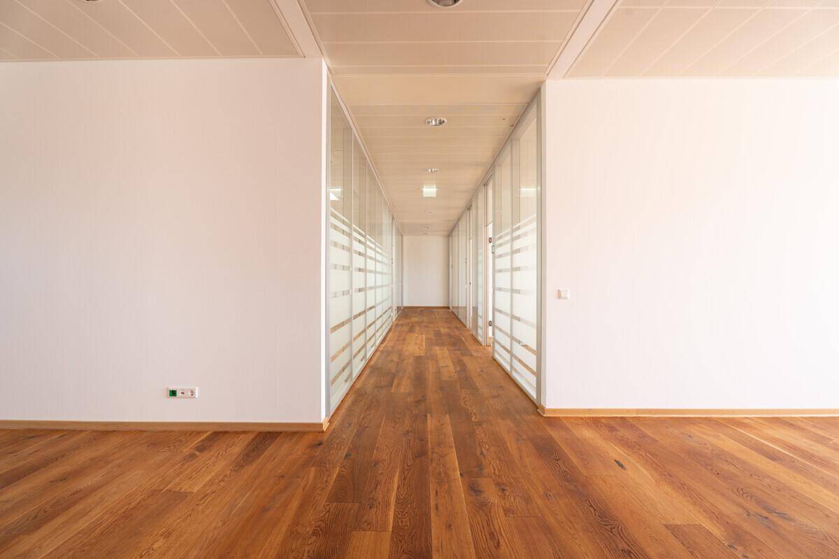 Langer Flur mit Holzboden und Glaswänden, die zu den Büroräumen führen.