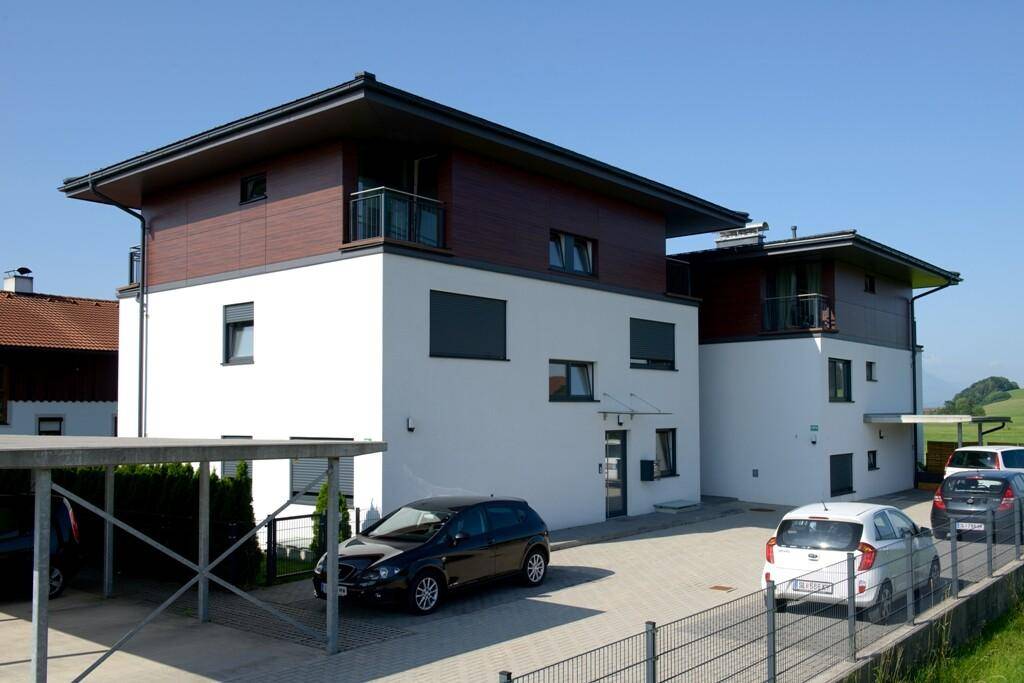 Zwei moderne Mehrfamilienhäuser mit weißer Fassade, Holzdetails und Carports auf gepflastertem Vorplatz.
