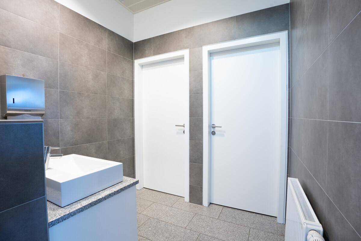 Moderner Toilettenraum mit grauen Fliesen, Waschbecken und zwei weißen Türen.