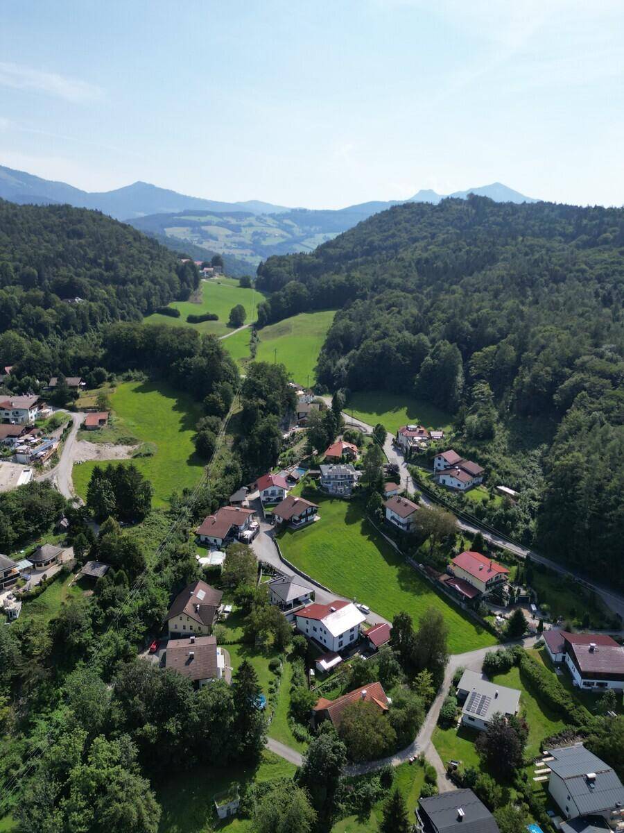 Idyllische Luftaufnahme einer Siedlung in einem Tal, umgeben von bewaldeten Hügeln und grünen Wiesen.