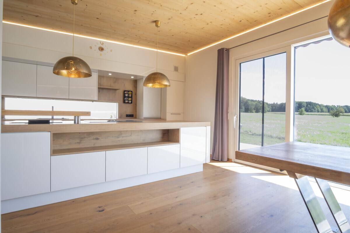 Moderne Küche mit weißen Schränken, Holztheke und großen Fenstern mit Blick auf die umliegende Natur.