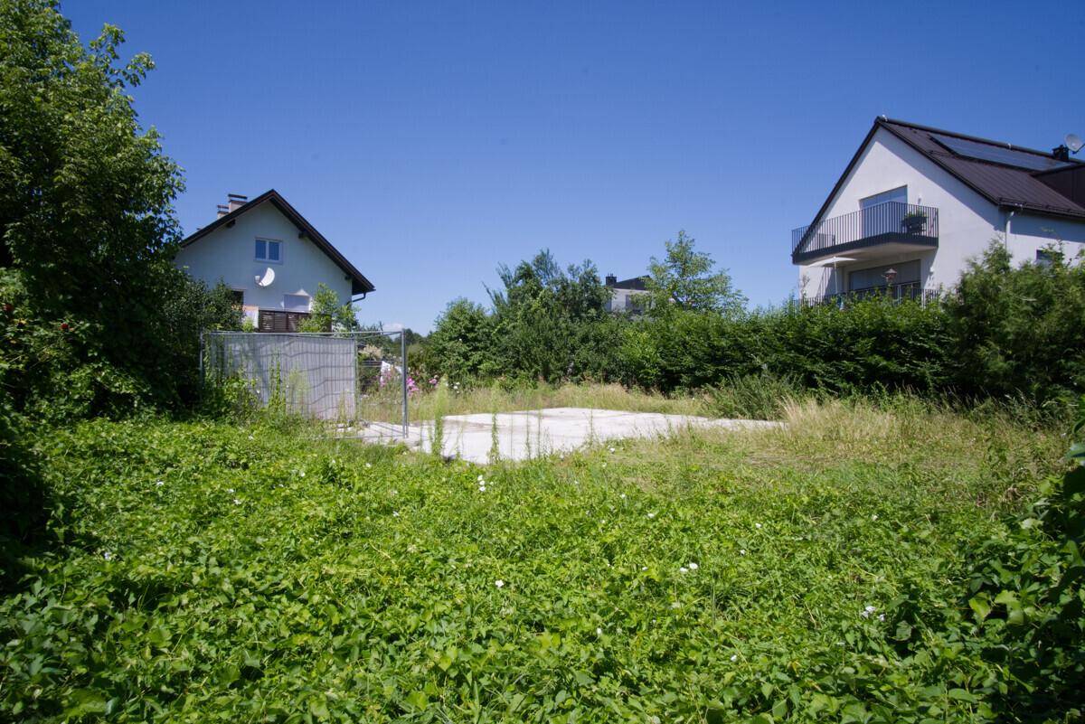 Unbebautes Grundstück mit hohem Gras und Blick auf benachbarte Wohnhäuser unter blauem Himmel.